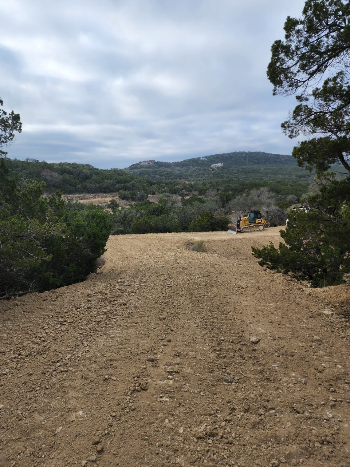 Graded dirt road and cleared pad with hills behind and a yellow dozer in the distance