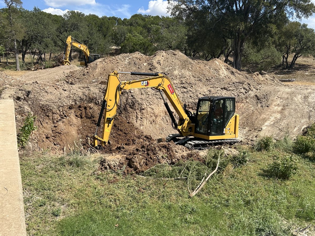 Excavator working in a cut beside a concrete wall, with soil piled behind it