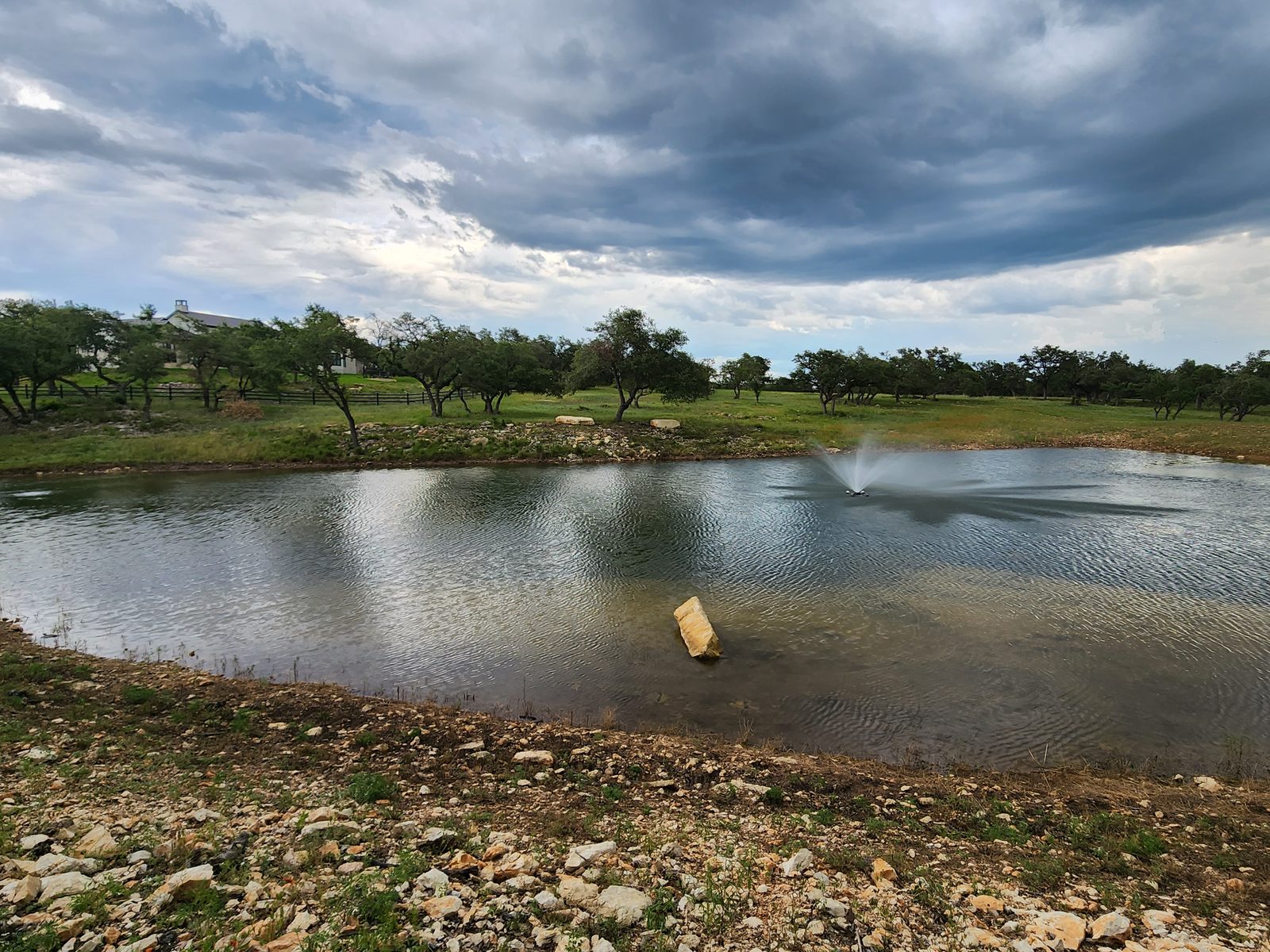Pond with a fountain aerator, a rock standing in the water and oaks along the far bank under a stormy sky