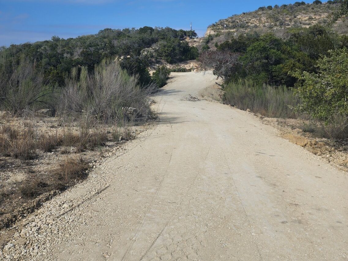 Pale caliche road running between hills, with brush along its edges